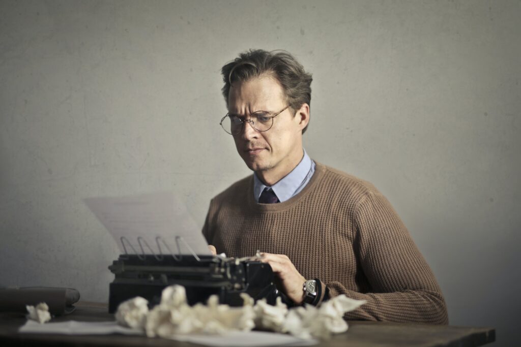 Man typing on a typewriter with many crumpled pieces of paper in the foreground. He's demonstrating that he's looking for creative writing jobs